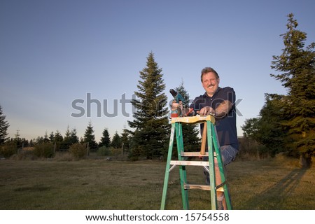 Man on ladder holding a drill and smiling. Horizontally framed photograph