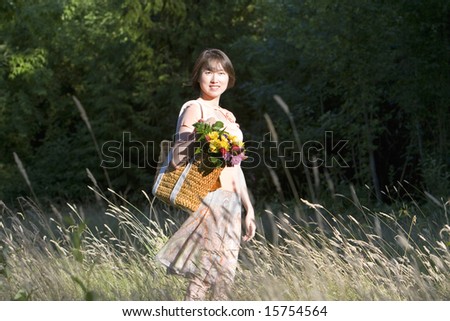 Attractive woman stands in a field while holding a basket containing a bouquet of flowers. She is standing to the side, smiling.  Vertically framed photo.