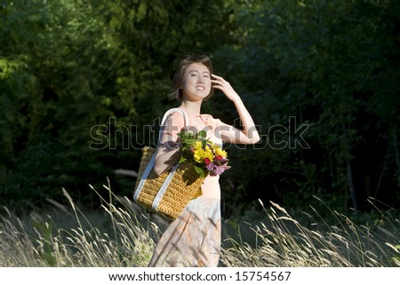Attractive woman stands in a field while holding a basket of flowers. She is standing to the side, brushing hair from her face.  Horizontally framed photo.