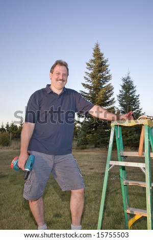 Man standing next to a ladder holding a drill and smiling. Vertically framed photograph