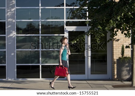 Young woman runs along building while talking on a cell phone. She is wearing a tank top and carrying a red bag. Horizontally framed photo.