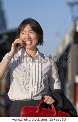 Young woman walks while talking on her cell phone. She is smiling and carrying a red bag. Vertically framed photo.