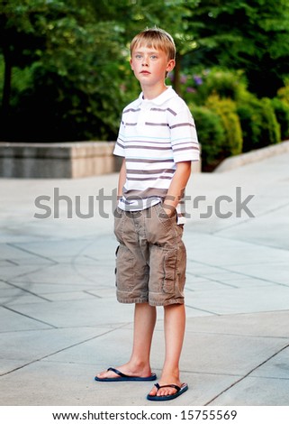 A young boy is standing outside on a sidewalk.  He is looking at the camera and has his hands in his pockets.  Vertically framed shot.