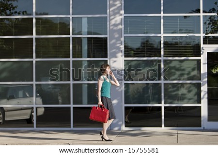 Young woman walks along building while talking on a cell phone and carrying a red bag. She is wearing a tank top. Horizontally framed photo.