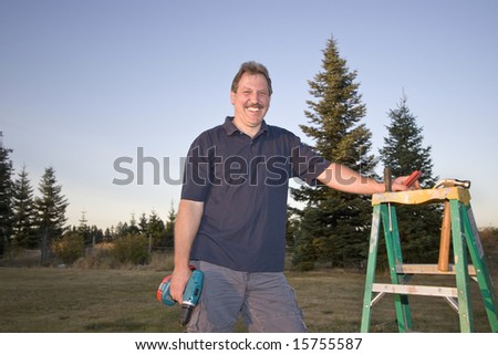 Man standing next to a ladder holding a drill and smiling. Horizontally framed photograph
