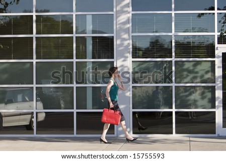 Young woman walks along building while talking on a cell phone and carrying a red bag. She is wearing a tank top. Horizontally framed photo.