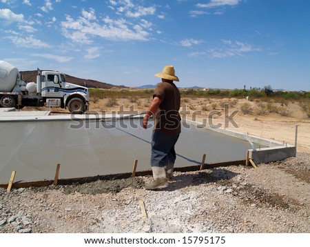 Construction worker smoothing out a freshly poured concrete slab. Shot is set in a desert with a white cement truck in the background. Horizontally framed shot.