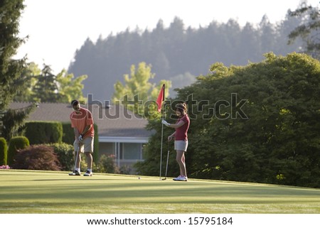 A young couple is setting up to play golf on the green of a golf course.  They are looking away from the camera.  Horizontally framed shot.
