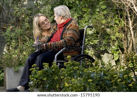 A younger woman is with her elderly mother in a garden.  She is smiling at the woman who is holding tablet and a pen, and looking back at her.   Horizontally framed shot.