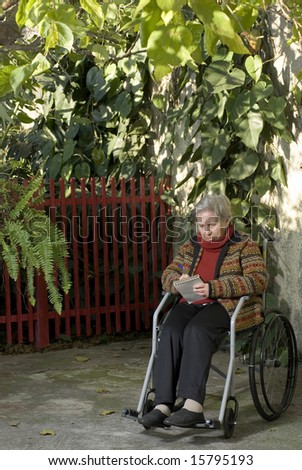 An elderly woman sitting in a wheelchair is in a garden.  She is writing something on a notepad and looking down at it.  Vertically framed shot.