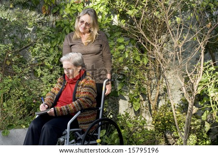 A younger woman is with her elderly mother in a garden.  She is smiling at the woman who is holding tablet and a pen, and looking down at what she is writing.   Horizontally framed shot.