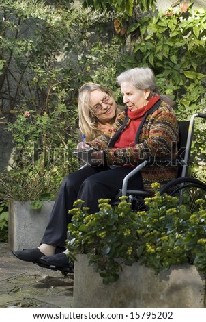 A younger woman is with her elderly mother in a garden.  The older woman is sitting in a wheelchair.  She is smiling at her mother who is looking away.  Vertically framed shot.
