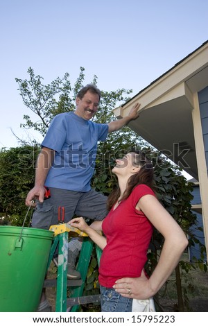 Man smiling standing on a ladder holding a bucket. His wife is standing next to him and they are laughing. Vertically framed photograph