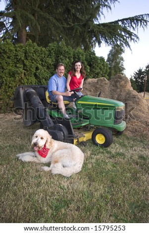 A man and woman sitting on a tractor smiling. Their dog is nearby on the grass. Vertically framed photograph
