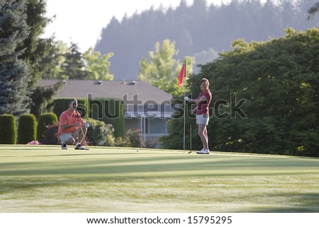 A young couple is setting up to play golf on the green of a golf course.  They are looking at the camera.  Horizontally framed shot.