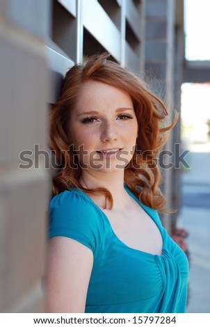 An attractive, young woman, half-smiles at the camera as she leans on a metal vent on a brick wall. Vertically framed shot.