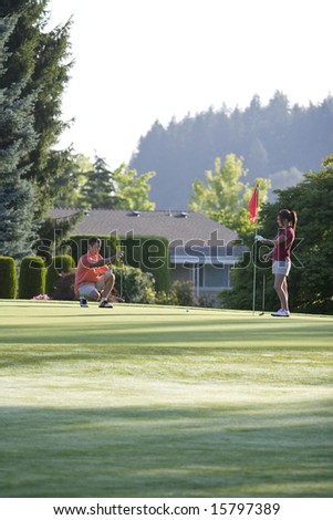 A young couple is setting up to play golf on the green of a golf course.  They are looking away from the camera.  Vertically framed shot.
