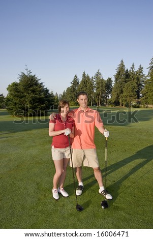 A young couple is standing on a golf course.  The man has his arms around the woman and they are both smiling at the camera.  Vertically framed shot.