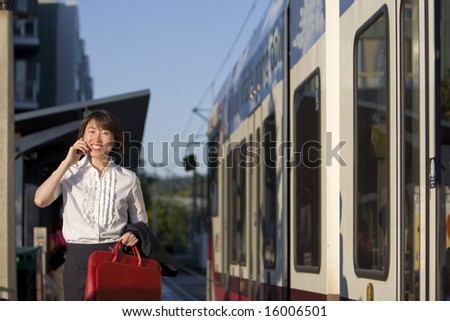 Smiling woman walks while talking on cell phone and holding red handbag. She is walking next to a train. Horizontally framed photo.