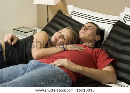 Man and woman relax on bed. Man has arm around woman while woman rest on his chest. Horizontally framed photo.