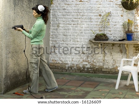 Young woman drills hole in wall. She is wearing a dust mask on her head. Horizontally framed photo.