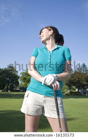 A young woman is standing on a golf course holding a club.  She is smiling, laughing, and looking away from the camera.  Vertically framed shot.