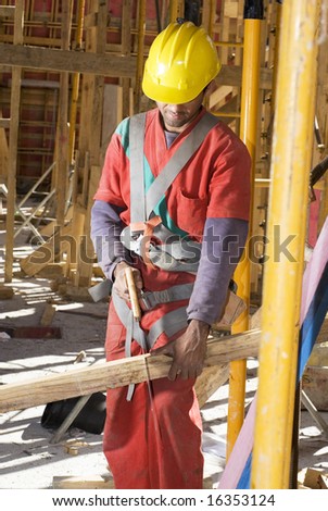 Construction worker stands while holding board. He is cutting it with a hand saw. Vertically framed photo.