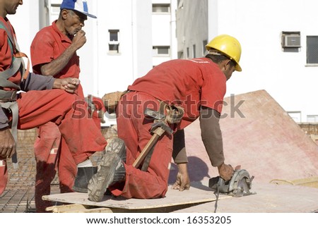 Construction worker cuts board with skill saw. He is wearing a tool belt and two other workers are watching him. Horizontally framed photo.