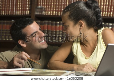 Couple smiles at each other in library. Man is kneeling and woman is sitting in front of laptop. Horizontally framed photo.