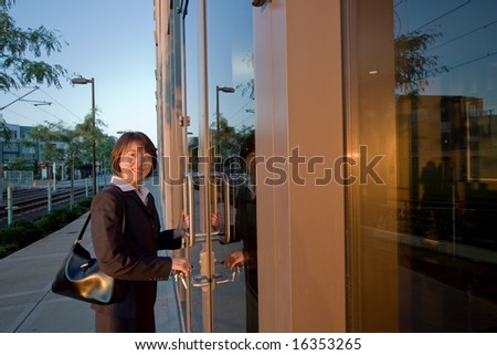 Woman wearing suit smiles at camera while opening door. She has a purse over her shoulder. Horizontally framed photo.