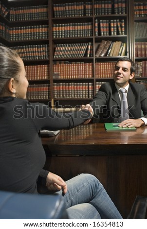 Man in suit sits behind desk. He shakes hand of woman on other side. Vertically framed photo.