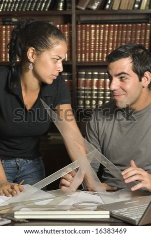Woman stands next to seated man at the library. They are looking at each other and holding rulers. Vertically framed photo.