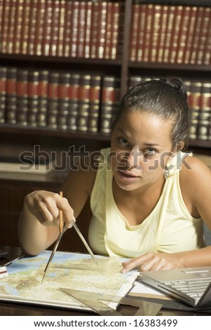 Woman uses compass to study map. She is sitting at a table in the library. Vertically framed photo.
