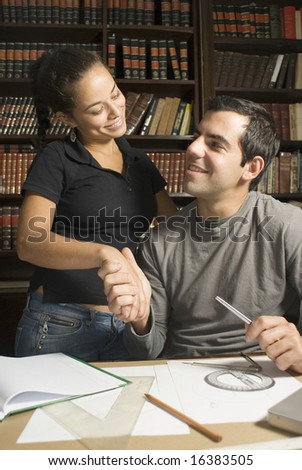 Seated man shakes hand of standing woman. They are studying and smiling at each other. Vertically framed photo.
