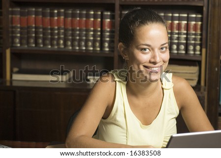 Woman sits at table in library. She is seated in front of her laptop smiling at the camera. Horizontally framed photo.