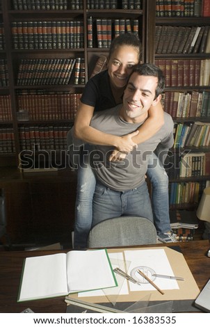 Woman rides on man\'s back in library. There are books on the table. Vertically framed photo.