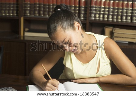 Woman smiles while writing in notebook. She is seated in a library. Horizontally framed photo.