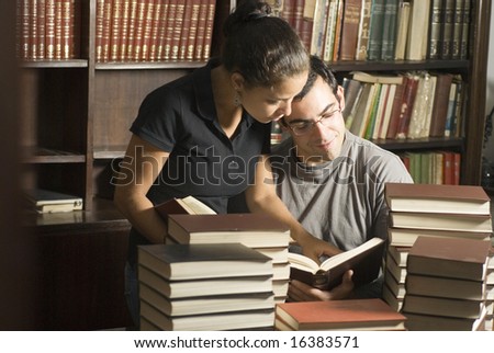 Woman stands next to man while they are studying at the library. She is pointing to a book while he is seated. Horizontally framed photo.