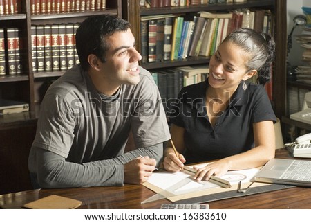 Couple studies in library. There are books and tools on the table and they are smiling. Horizontally framed photo.