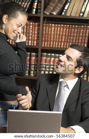 Man and woman shake hands over laptop. They are smiling at each other while she is on the cellphone. Vertically framed photo.