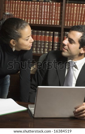 Man sitting at table in library looks at woman standing next to him. They are wearing suits and looking at each other. Vertically framed photo.