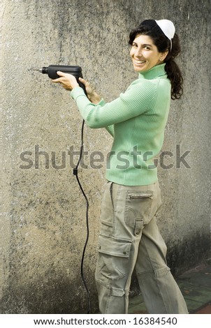 Woman drills hole in wall. She is smiling and wearing a dust mask on her head. Vertically framed photo.