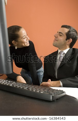 Businessman and woman smiling at each other. He is seated at his desk by his computer. Vertically framed photo.