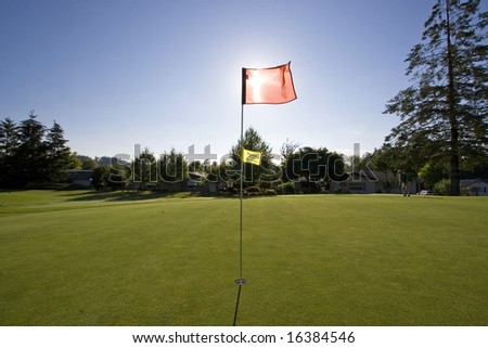 View of a golf course as the sun is going down.  The flag is blocking the sun.  Horizontally framed shot.