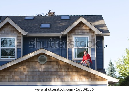 A man, sitting on top of his roof, holds a power drill, in an attempt to fix his roof, staring at the camera, smiling. Horizontally framed shot.