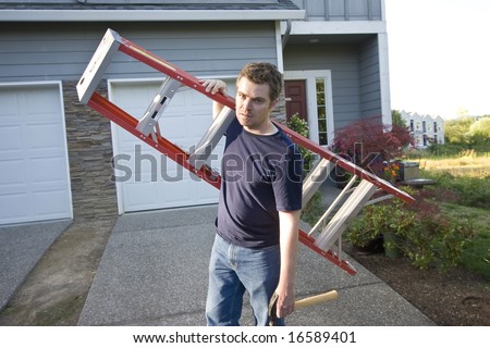 Angry, frowning man standing in front of house holding ladder and hammer. Horizontally framed photo.