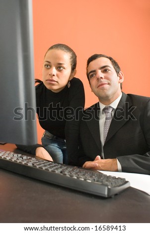 Businessman sits at his desk as his co-worker stands behind him and they look at a computer. Vertically framed photo.