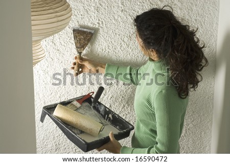 Woman holds a scraper and scrapes wall while holding paint tray and roller. Vertically framed photo.