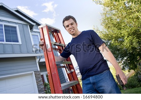 Unhappy man standing in front of house holding ladder and hammer. Horizontally framed photo.