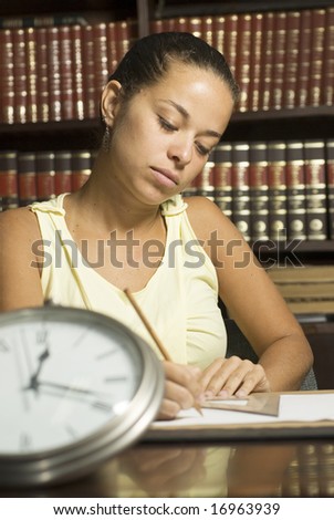 Student working while seated at a desk in an office surrounded by books and a clock. Vertically framed photo.